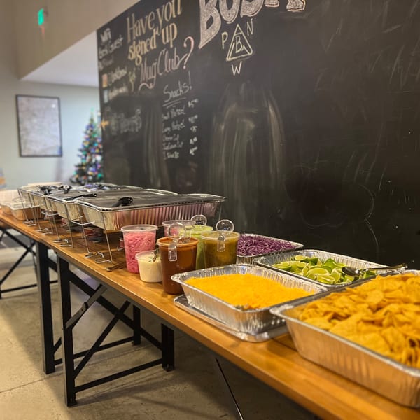 A buffet line with various Mexican food items, including hot trays, salsas, shredded cabbage, limes, and tortilla chips.