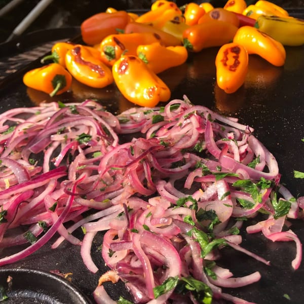 Sautéing vibrant red onions with green herbs and small orange peppers on a dark griddle.